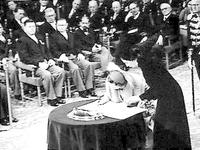 Koningin Juliana ondertekent in de Ridderzaal het Statuut. Naast haar staat mevrouw Tellegen, directeur van het Kabinet der Koningin.