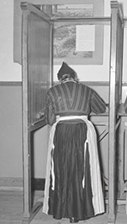 Een zwart-wit foto van een mevrouw in een stemhok die het stembiljet invult.